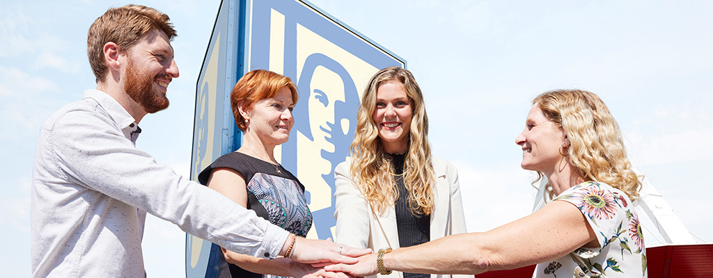 Vier Personen stehen vor einem großen ROLAND Wappen. Sie halten jeweils eine Hand in die Mitte und haben diese übereinander liegen. Alle vier lächeln.