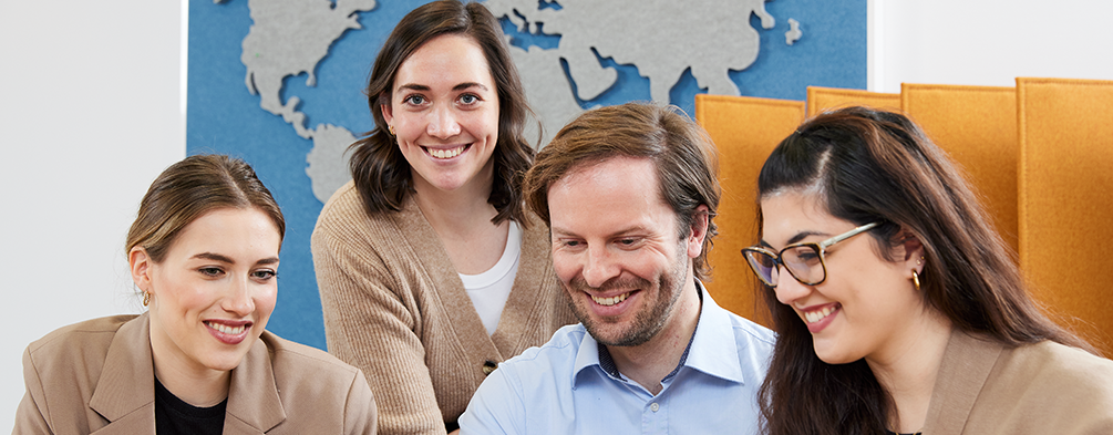 Junge Frau steht vor einem Tisch und schaut nach vorn. An dem Tisch sitzen 3 weitere junge Menschen und schauen gemeinsam auf einen Laptop. Alle lächeln.