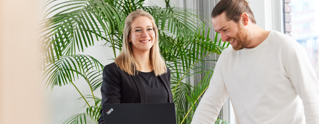 Junge Frau schaut nach vorne und lächelt. Neben ihr steht ein Mann. Im Hintergrund ist ein Fenster.
