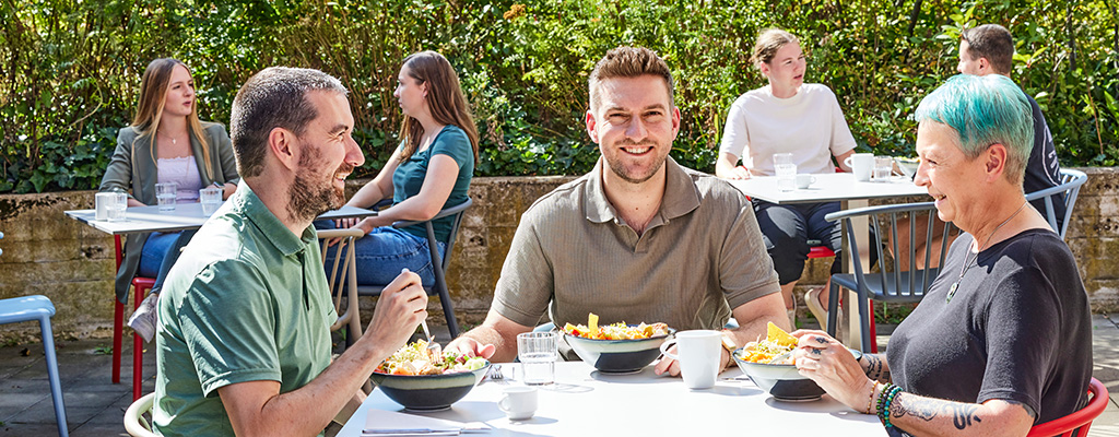 Drei Personen sitzen draußen an einem Tisch. Sie haben vor sich Schalen mit Essen stehen. Alle lächeln. Hinter ihnen sitzen weitere Personen an Tischen.