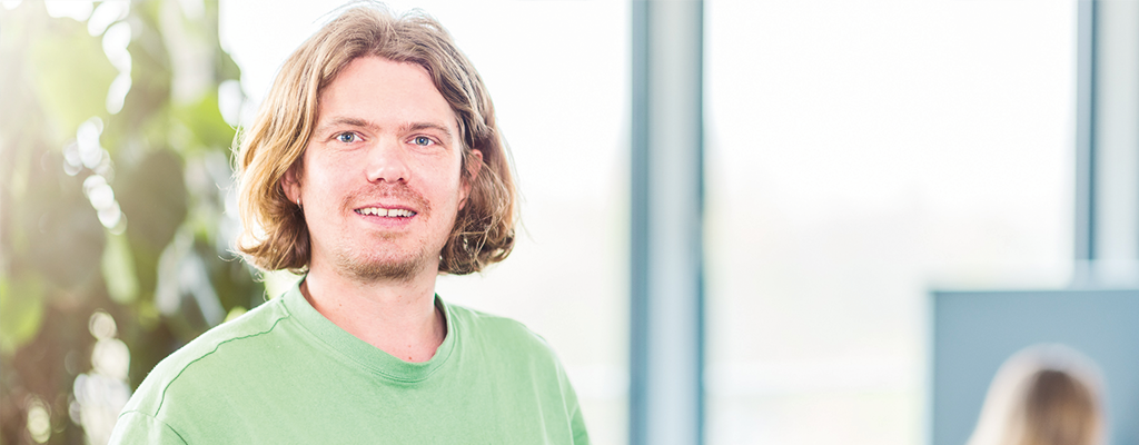 Junger Mann steht in einem Büroraum. Er schaut nach vorne und lächelt.