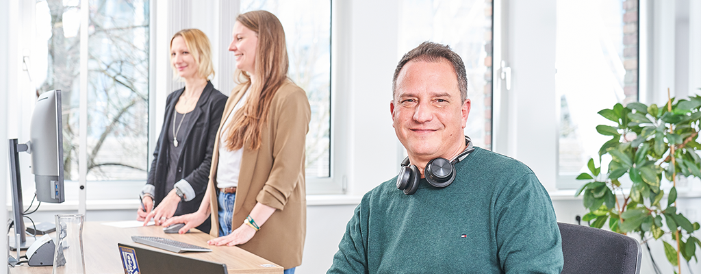 Mann sitzt an einem Schreibtisch. Er hat ein Headset um den Hals, schaut nach vorn und lächelt. Neben ihm stehen zwei Frauen an einem weiteren Schreibtisch und schauen auf dem Bildschirm. Sie lächeln.
