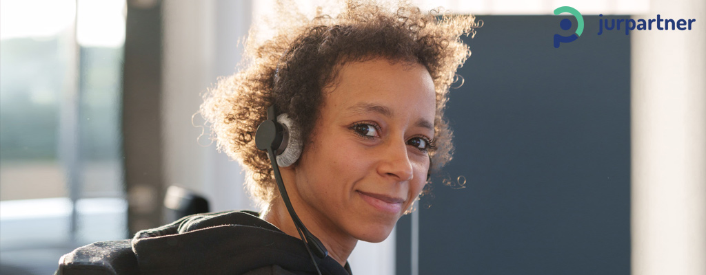 Junge Frau mit Headset schaut nach vorn und lächelt. Dahinter ist ein Büroraum.