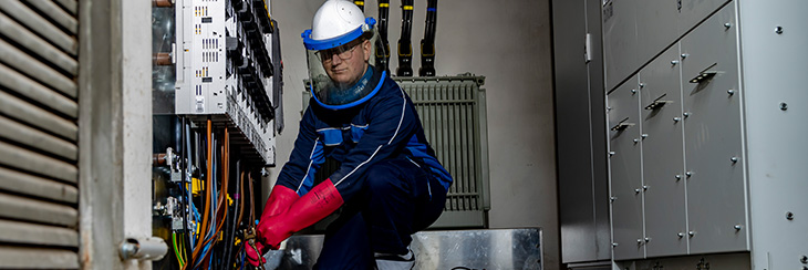 Ein Elektromonteur kniet mit Schutzausrüstung vor den Kabeln eines Schaltschranks